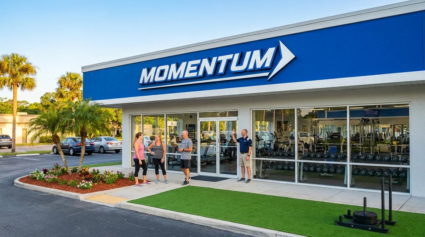 Momentum Fitness and Wellness Center: a clean, welcoming local gym with fresh landscaping, members chatting at the door, and a sled on the turf